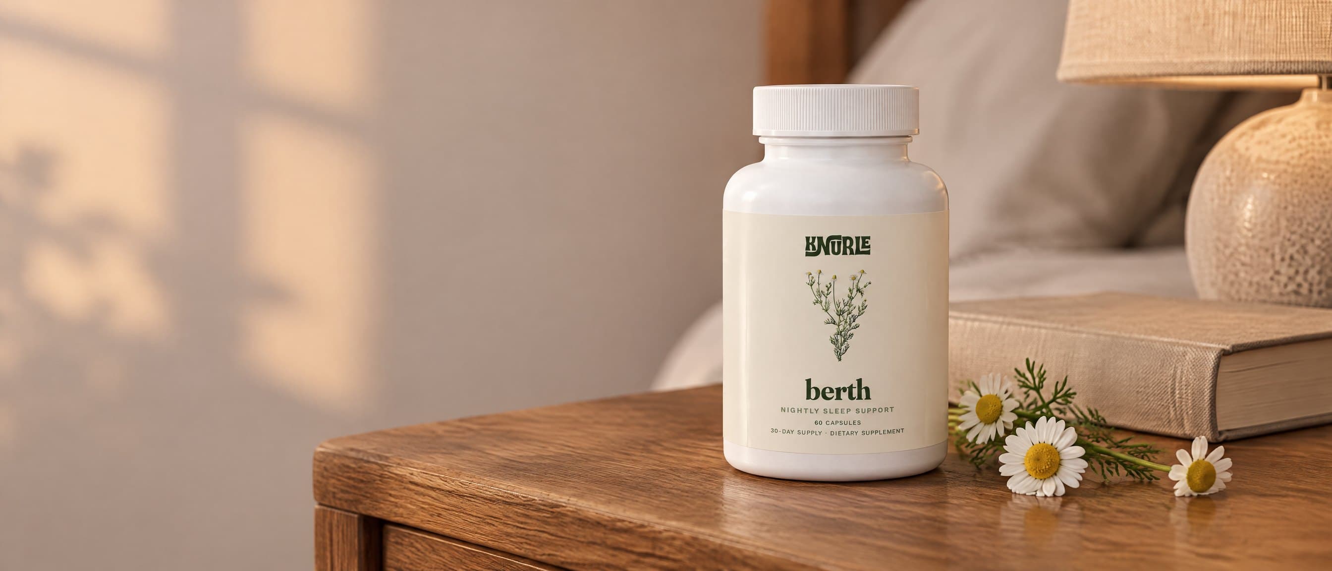 A bottle of Berth on a wooden nightstand beside a book and fresh chamomile flowers