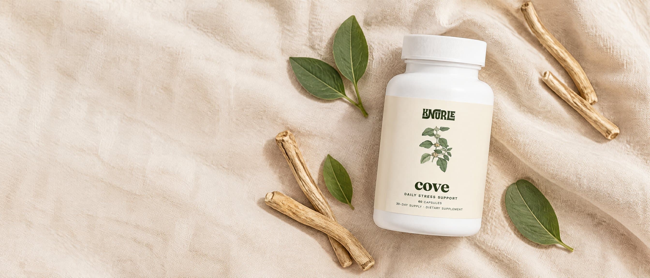 A bottle of Cove photographed from above on cream linen among ashwagandha roots and green leaves